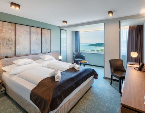 Modernes Hotelzimmer mit zwei Betten, Schreibtisch, Stuhl und Fenster mit Blick auf das Meer und die ferne Küste.