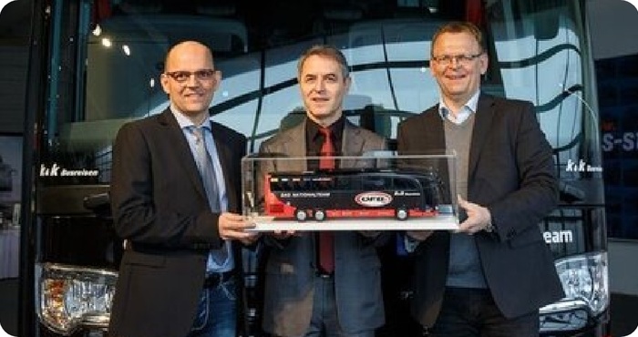 Drei Männer in Anzügen, die lächelnd einen Modellbus vor einen großen schwarzen Bus halten.