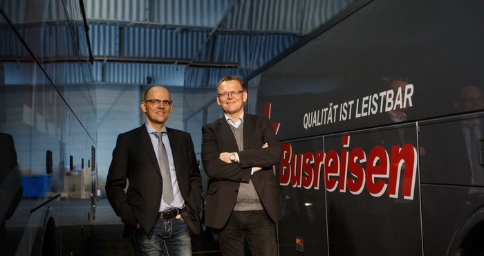 Zwei Männer in Geschäftskleidung stehen neben einem Bus mit der Aufschrift Busreisen in einer Garage.
