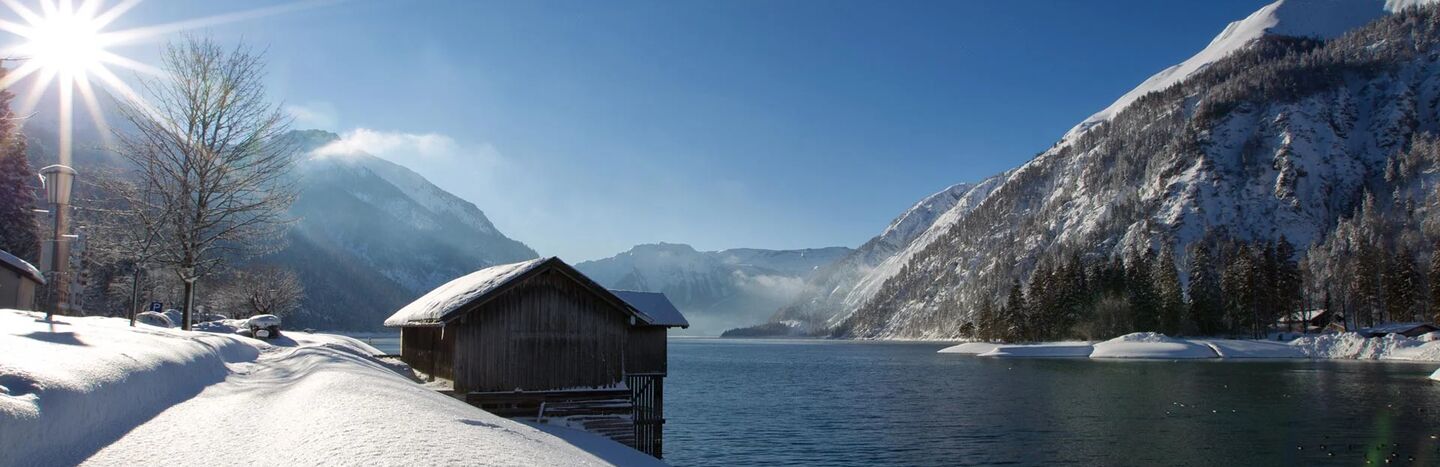 Eine Holzhütte an einem verschneiten Seeufer, mit Bergen im Hintergrund unter einem klaren blauen Himmel.