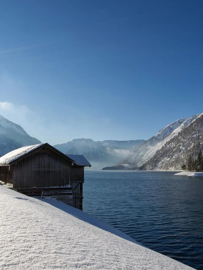 Eine Holzhütte an einem verschneiten Seeufer, mit Bergen im Hintergrund unter einem klaren blauen Himmel.