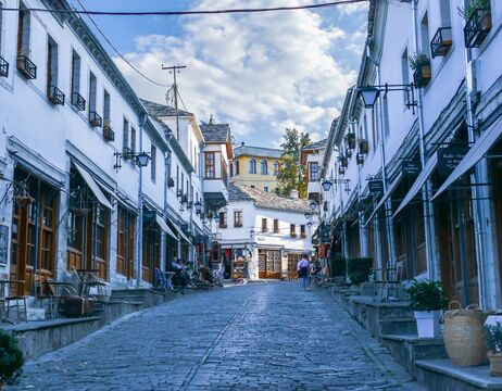 Eine Straße mit Kopfsteinpflaster, gesäumt von weißen Gebäuden und Geschäften unter einem teilweise bewölkten Himmel.
