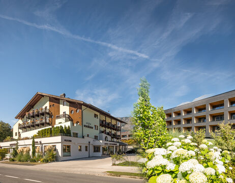 Ein Hotel mit Balkonen und Blumen davor, neben einem modernen Gebäude, unter einem blauen Himmel mit Wolkenfetzen.