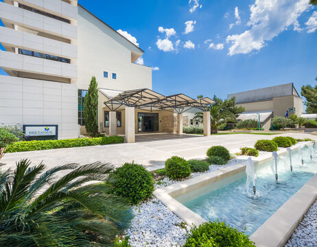 Moderner Gebäudeeingang mit Glasvordach, Landschaftsgarten und kleinem Springbrunnen unter strahlend blauem Himmel.