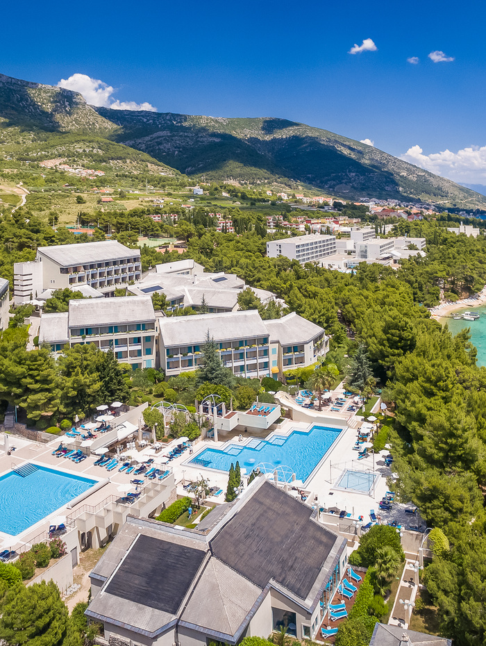 Luftaufnahme einer Anlage mit Pools, umgeben von Bäumen, in der Nähe der Küste und der Berge unter einem klaren blauen Himmel.