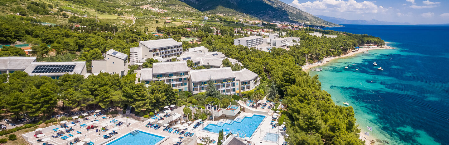 Luftaufnahme einer Anlage mit Pools, umgeben von Bäumen, in der Nähe der Küste und der Berge unter einem klaren blauen Himmel.