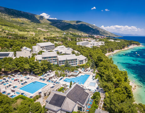 Luftaufnahme einer Anlage mit Pools, umgeben von Bäumen, in der Nähe der Küste und der Berge unter einem klaren blauen Himmel.
