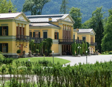 Gelbe Villa mit grünen Fensterläden, Säulen und Efeu, umgeben von Gärten und Bergen im Hintergrund.