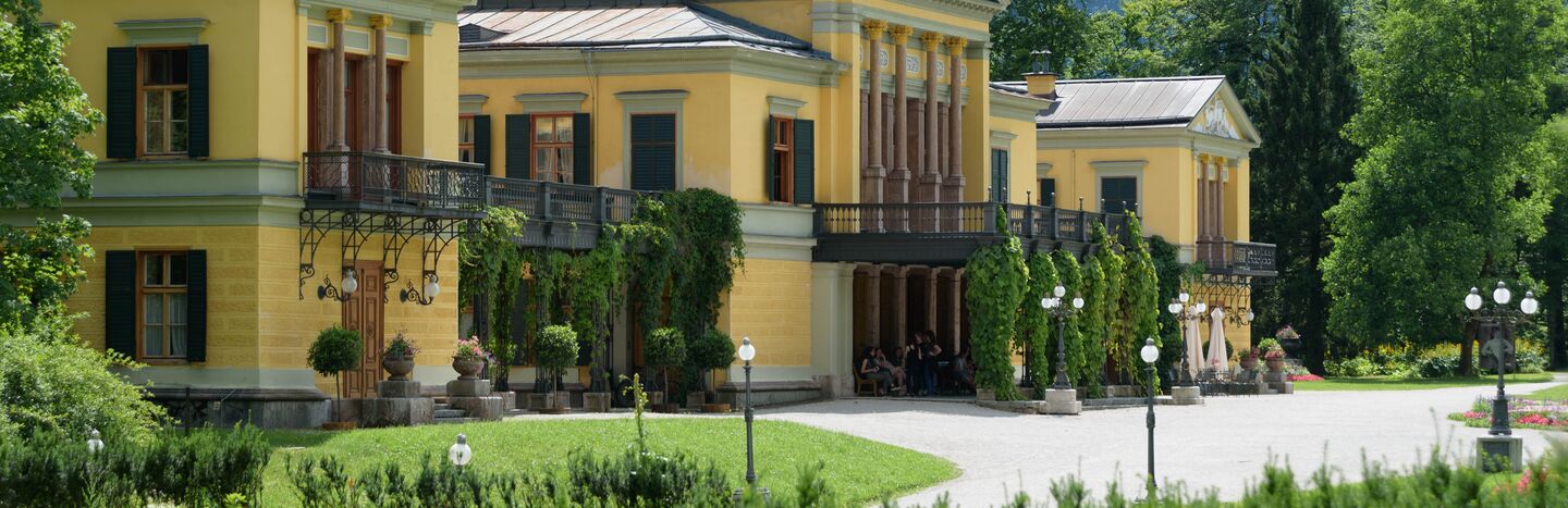 Gelbe Villa mit grünen Fensterläden, Säulen und Efeu, umgeben von Gärten und Bergen im Hintergrund.