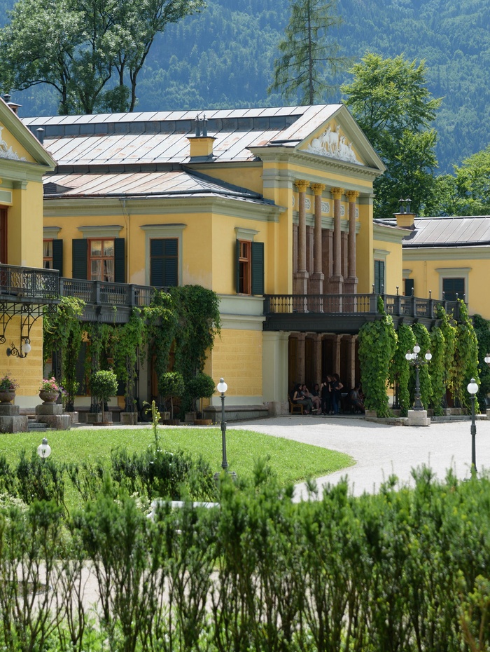 Gelbe Villa mit grünen Fensterläden, Säulen und Efeu, umgeben von Gärten und Bergen im Hintergrund.