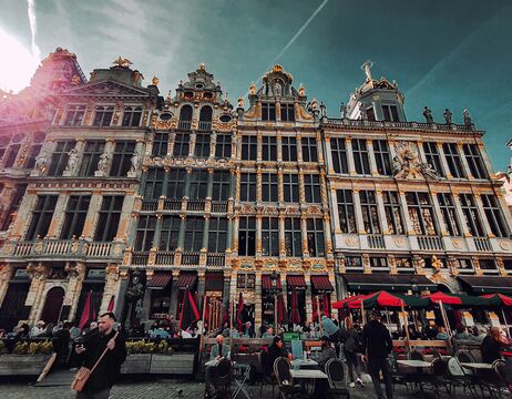 Verschnörkelte historische Gebäude mit goldenen Akzenten und Straßencafés unter roten Sonnenschirmen auf einem belebten Platz.