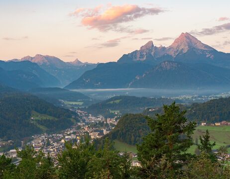 Berglandschaft bei Sonnenaufgang mit einer Stadt in einem grünen Tal und Wolken über entfernten Gipfeln.