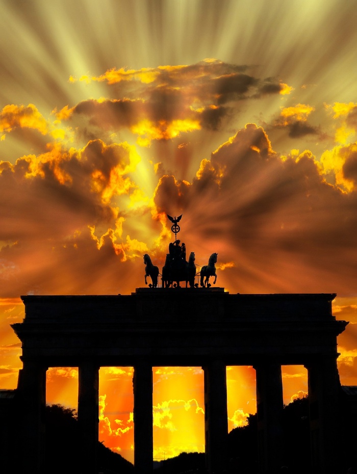 Silhouette des Brandenburger Tors bei Sonnenuntergang mit dramatischen Sonnenstrahlen, die durch die Wolken brechen.