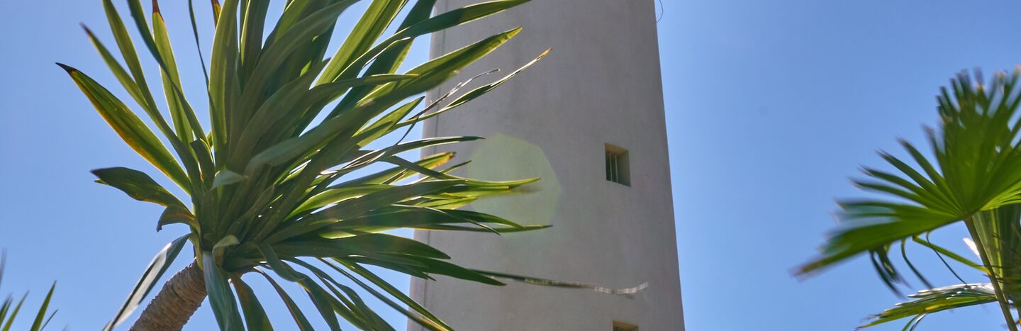 Ein hoher weißer Leuchtturm mit Palmen und strahlender Sonne vor einem klaren blauen Himmel.