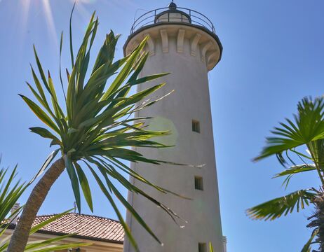 Ein hoher weißer Leuchtturm mit Palmen und strahlender Sonne vor einem klaren blauen Himmel.