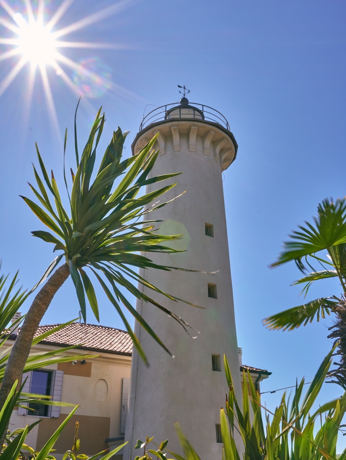 Ein hoher weißer Leuchtturm mit Palmen und strahlender Sonne vor einem klaren blauen Himmel.