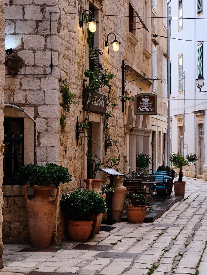 Schmale Kopfsteinpflasterstraße mit Topfpflanzen, rustikalen Steingebäuden und Straßencafés.