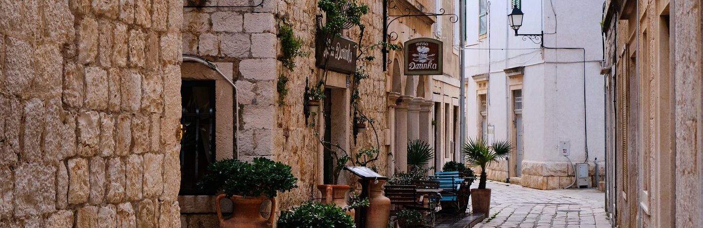 Schmale Kopfsteinpflasterstraße mit Topfpflanzen, rustikalen Steingebäuden und Straßencafés.