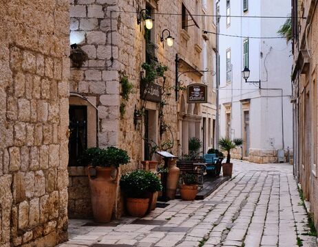 Schmale Kopfsteinpflasterstraße mit Topfpflanzen, rustikalen Steingebäuden und Straßencafés.