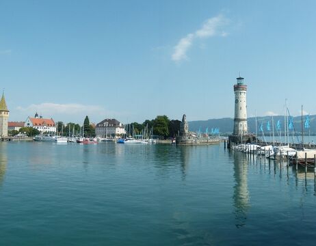Hafen mit Segelbooten, einem Leuchtturm und einer Löwenstatue bei ruhigem Wasser und strahlend blauem Himmel.