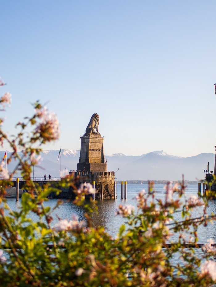 Eine steinerne Löwenstatue steht auf einer Hafenmole, eingerahmt von rosa Blumen und Bergen im Hintergrund.