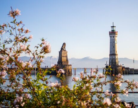 Eine steinerne Löwenstatue steht auf einer Hafenmole, eingerahmt von rosa Blumen und Bergen im Hintergrund.
