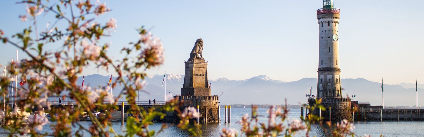 Eine steinerne Löwenstatue steht auf einer Hafenmole, eingerahmt von rosa Blumen und Bergen im Hintergrund.