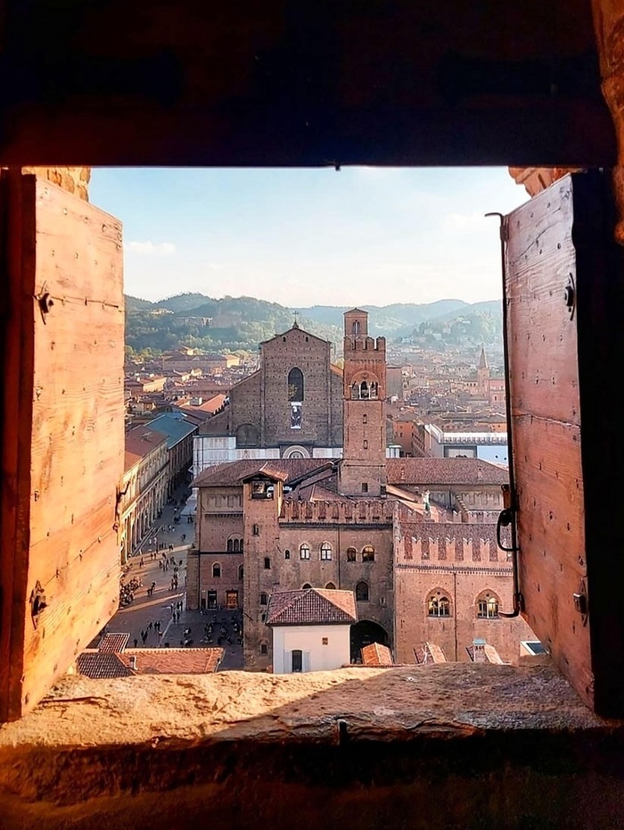 Blick auf historische Gebäude und weit entfernte Hügel in Bologna, Italien, durch ein offenes Holzfenster gesehen.