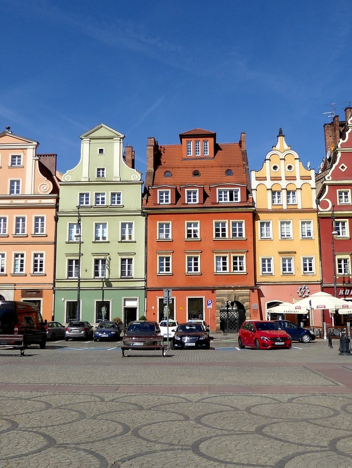 Bunte historische Gebäude mit verzierten Fassaden und parkende Autos auf einem sonnigen europäischen Stadtplatz.
