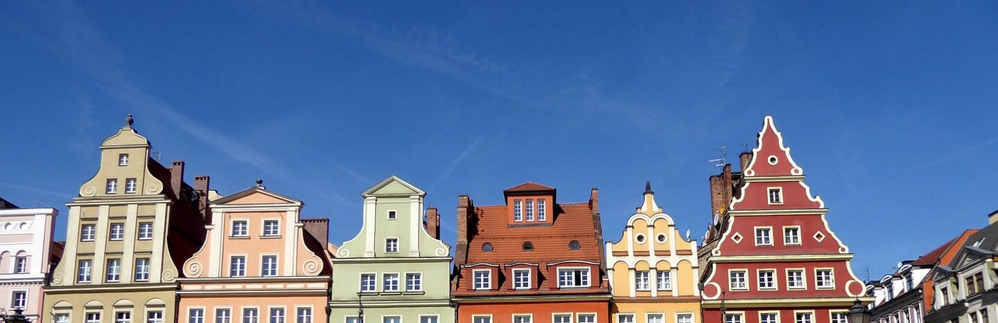 Bunte historische Gebäude mit verzierten Fassaden und parkende Autos auf einem sonnigen europäischen Stadtplatz.