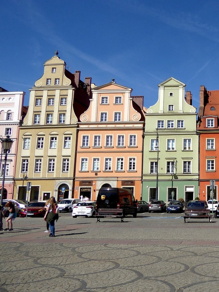 Bunte historische Gebäude mit verzierten Fassaden und parkende Autos auf einem sonnigen europäischen Stadtplatz.