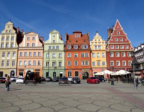 Bunte historische Gebäude mit verzierten Fassaden und parkende Autos auf einem sonnigen europäischen Stadtplatz.