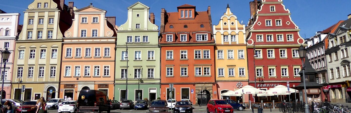 Bunte historische Gebäude mit verzierten Fassaden und parkende Autos auf einem sonnigen europäischen Stadtplatz.