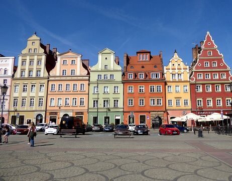 Bunte historische Gebäude mit verzierten Fassaden und parkende Autos auf einem sonnigen europäischen Stadtplatz.
