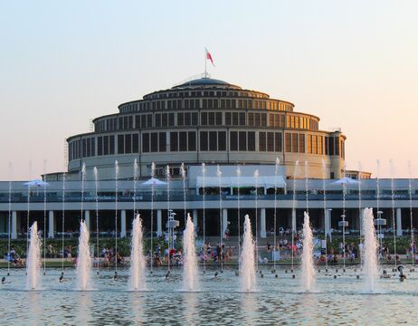Rundes Gebäude mit vielen Fenstern hinter einem großen Springbrunnen, unter einem klaren Himmel bei Sonnenuntergang.