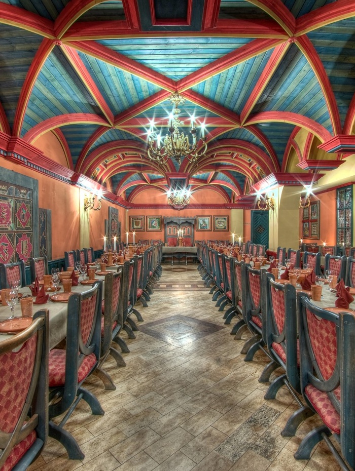 Langer Speisesaal mit farbenfroher Gewölbedecke, roten Stühlen, gedeckten Tischen und verzierten Kronleuchtern.