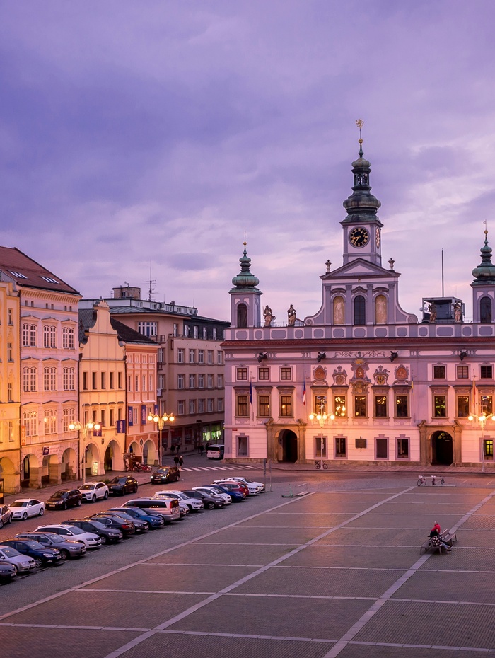 Stadtplatz in der Abenddämmerung mit geparkten Autos und einem historischen Gebäude mit Uhrenturm und Turmspitzen, die unter dem violetten Himmel leuchten.
