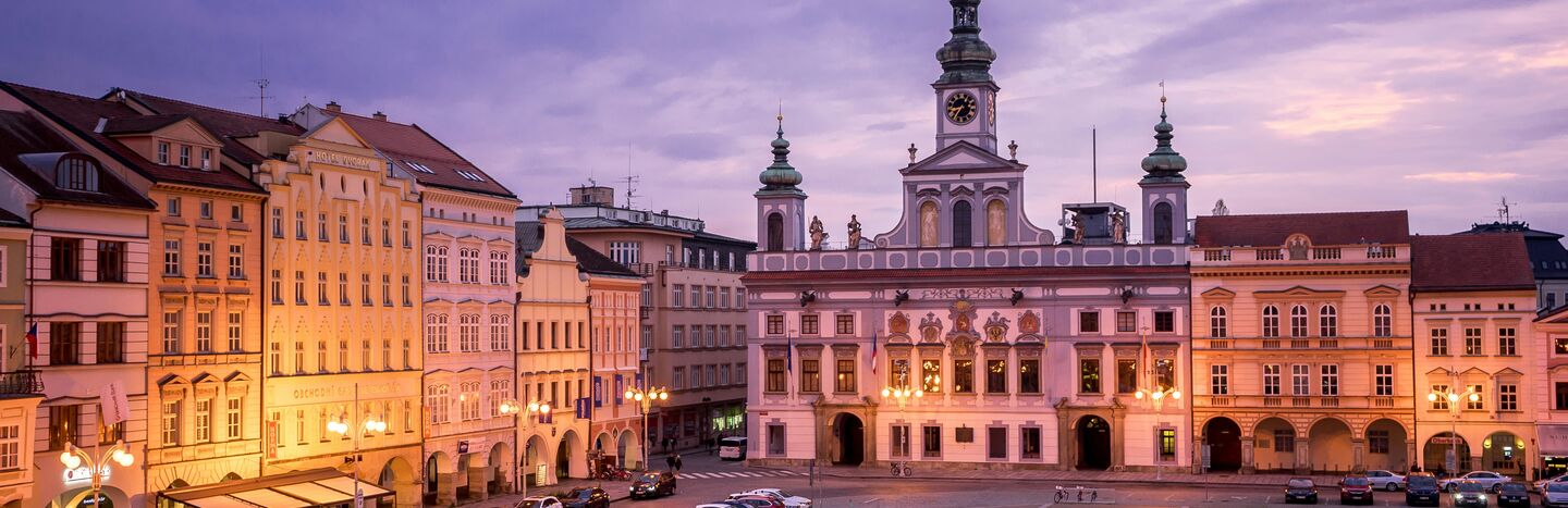 Stadtplatz in der Abenddämmerung mit geparkten Autos und einem historischen Gebäude mit Uhrenturm und Turmspitzen, die unter dem violetten Himmel leuchten.