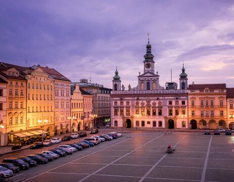 Stadtplatz in der Abenddämmerung mit geparkten Autos und einem historischen Gebäude mit Uhrenturm und Turmspitzen, die unter dem violetten Himmel leuchten.