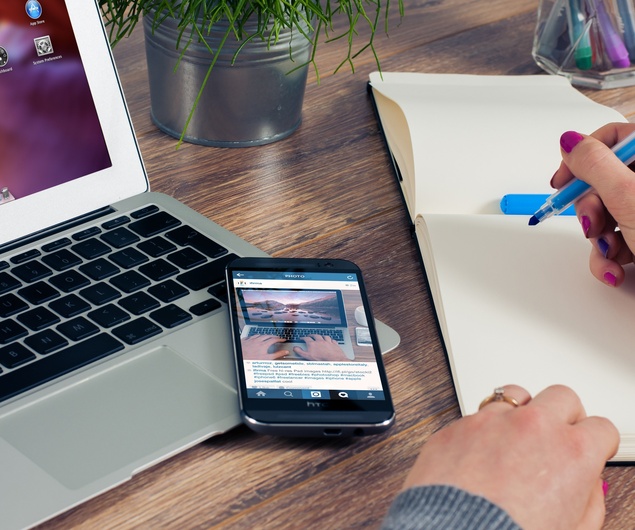 Person, die in ein Notizbuch schreibt, neben einem Laptop und einem Smartphone auf einem Holztisch mit einer Pflanze im Hintergrund.