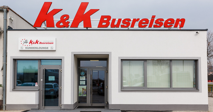 Weißes Gebäude mit großem roten K & K Busreisen-Schild und Glastüren, tagsüber von vorne gesehen.