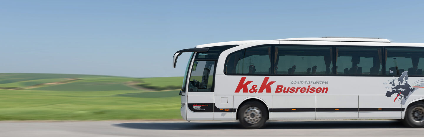 Ein weißer Bus mit der Aufschrift K&K Busreisen fährt auf einer Straße durch eine grüne, hügelige Landschaft.