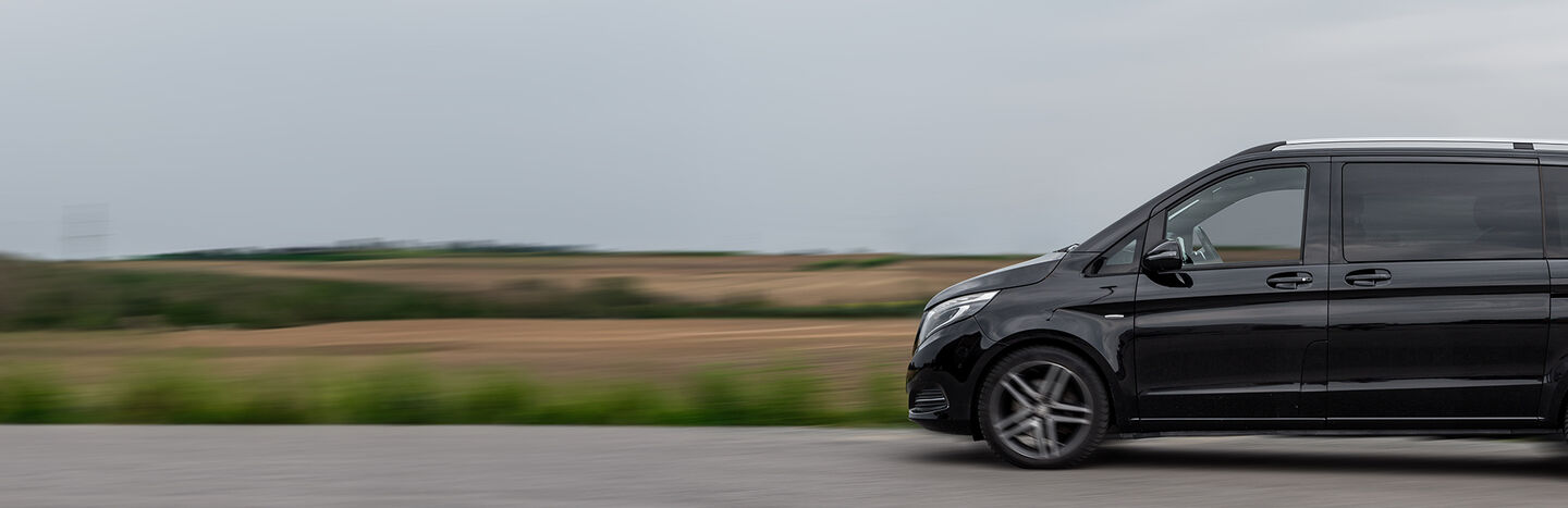 Ein schwarzer Lieferwagen fährt auf einer Straße mit verschwommenen Feldern und Himmel im Hintergrund.