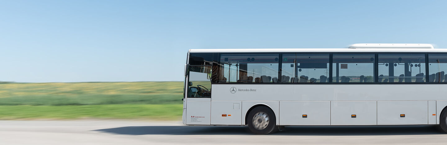Ein weißer Mercedes-Benz-Bus fährt auf einer Straße mit grünen Feldern im Hintergrund unter einem klaren Himmel.
