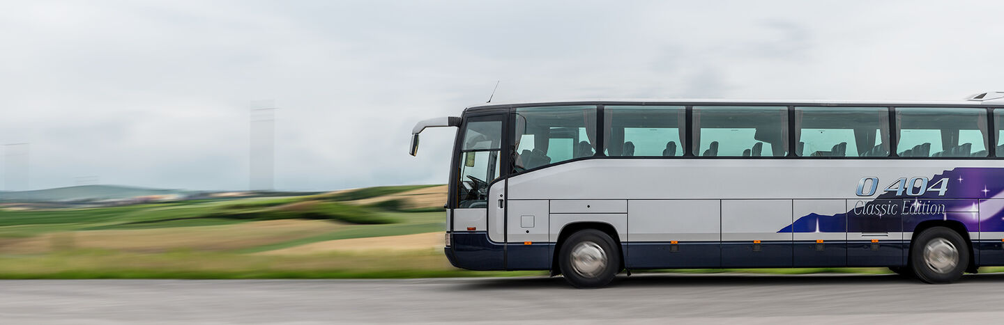 Ein weißer und lilafarbener Reisebus fährt auf einer Straße mit verschwommenen grünen Feldern im Hintergrund.