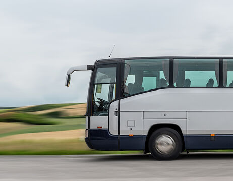 Ein weißer und lilafarbener Reisebus fährt auf einer Straße mit verschwommenen grünen Feldern im Hintergrund.