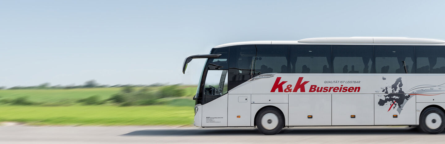 Weißer Reisebus mit k&k Busreisen-Logo fährt auf einer Straße mit grünen Feldern im Hintergrund.
