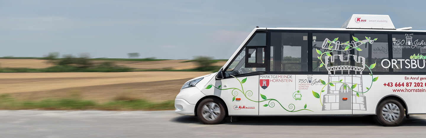 Weißer Shuttlebus mit grüner Rankengrafik, der auf einer Landstraße unter blauem Himmel an Feldern vorbeifährt.