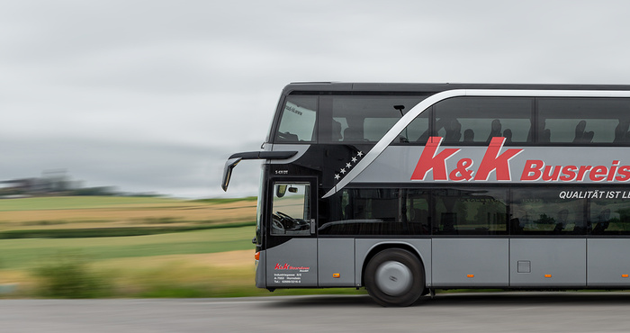 Ein doppelstöckiger K&K-Reisebus fährt unter einem bewölkten Himmel an grünen Feldern vorbei.