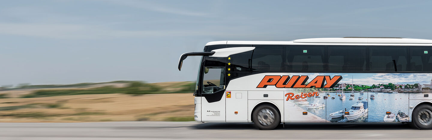 Ein weißer Reisebus mit Pulay Reisen und einer Hafenszene fährt auf einer Straße mit verschwommener Landschaft.